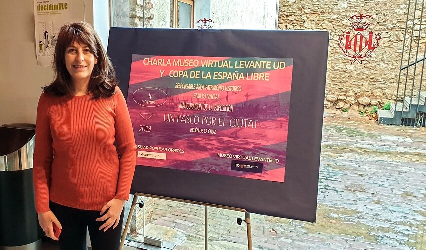 Una mujer de pie en el interior junto a un gran cartel para una charla y un acto virtuales en un museo, con muros de piedra y un patio visibles al fondo.