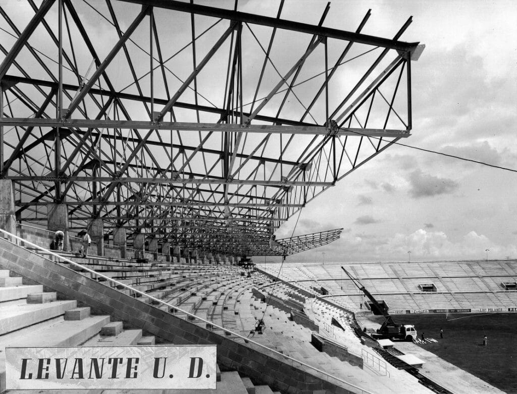 Gradas del estadio parcialmente construidas con estructura de acero vista y asientos vacíos. Un cartel indica "Levante U.D." y se ven vehículos de construcción en el campo.