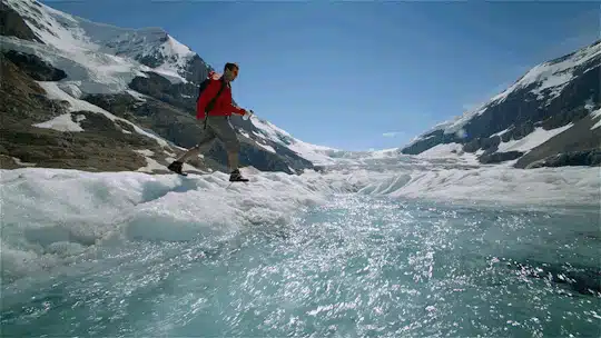 Una persona con chaqueta roja camina por el borde de un glaciar junto a un arroyo de hielo derretido, rodeado de montañas nevadas bajo un cielo azul despejado.