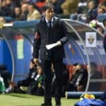 Un hombre con traje y abrigo se sitúa en la banda de un campo de fútbol, sosteniendo unos papeles, con los jugadores y los suplentes sentados detrás de él en un banquillo.
